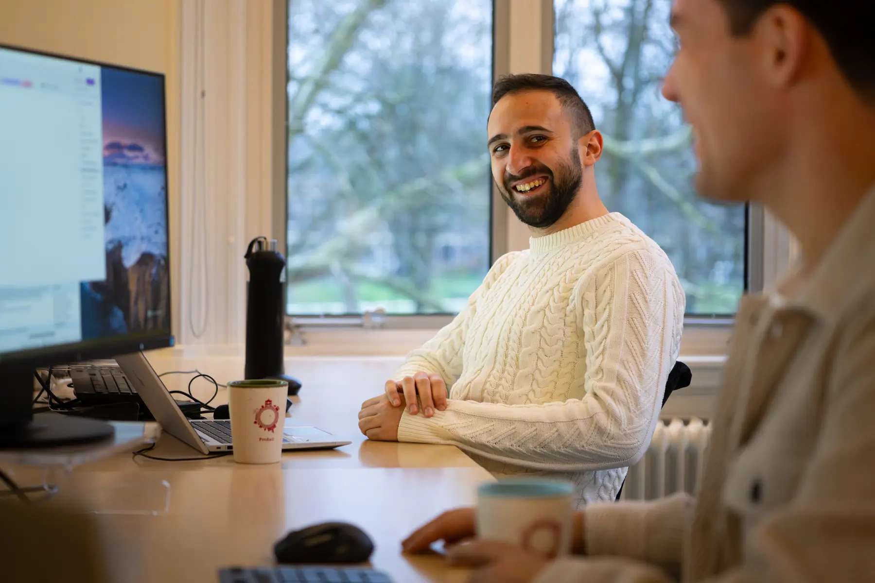 Madj zit aan zijn bureau achter zijn computer in gesprek met een collega links van hem.